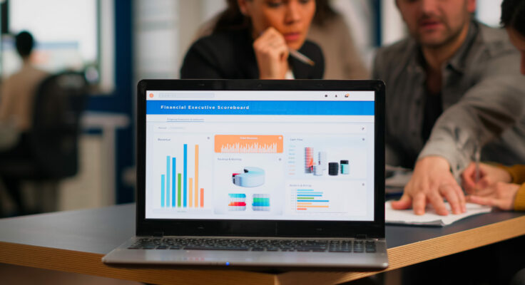 Laptop screen ready for financial project presentation placed on desk while business people work in background. Diverse employees gathered in co-working, working process in busy company, teamwork help